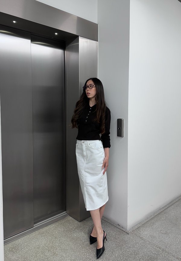Donna con i capelli lunghi che indossa occhiali, top nero, gonna bianca e tacchi neri, in piedi vicino alle porte chiuse dell'ascensore in un ambiente interno moderno.