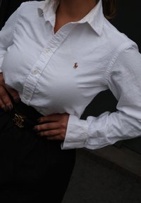 Camicia bianca con colletto e piccolo logo polo arancione, indossata con una cintura nera con fibbia dorata con le lettere "RL". La trama appare liscia e nitida.