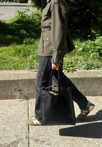 Black leather tote bag with a smooth texture and no visible hardware. Carried by a person wearing an olive-green jacket and black jeans.