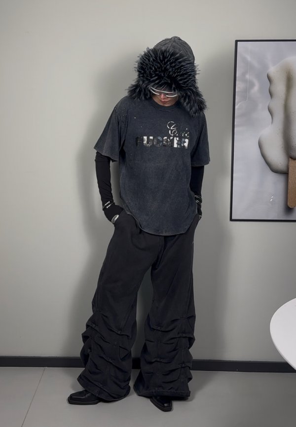 T-shirt nero oversize con texture vissuta e testo argentato, abbinato a pantaloni neri larghi caratterizzati da dettagli di tessuto arricciato e un cappuccio in pelliccia.