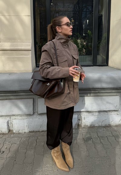 Chaqueta de utilidad marrón con múltiples bolsillos, pantalones negros de pierna ancha, botas de vaquero color beige con costuras decorativas; sosteniendo una taza de bebida helada.