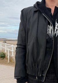 Crna bomber jakna glatke završne obrade, s elastičnim pojasom i zatvaračem na patent, slojevito nošena preko tamne grafičke hoodice.