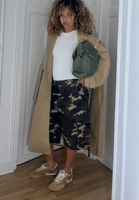 Beżowy płaszcz trencz na białej koszulce, w połączeniu z moro szortami cargo i beżowymi trampkami. Trzyma zieloną torbę. Tło z drewnianą podłogą.