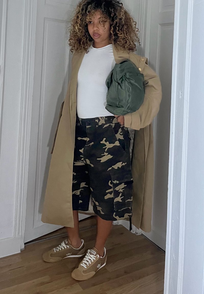 Bež plašč tipa trench čez belo majico, kombiniran s kratkimi hlačami s kamuflažno vzorcem in bež supergami. Drži zeleno torbo. Ozadje lesenega poda.