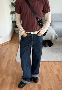 Άτομο που φοράει στρωμένα μακριά μπεζ μανίκια κάτω από μακρυμάνικη μπλούζα μπορντό, παντελόνι με φαρδιά μπατζάκια και μανσέτες, μαύρα παπούτσια και μαύρη τσάντα χιαστί, σε εσωτερικό χώρο.