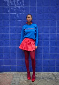 Sweatshirt azul com o logo "LACOSTE" em vermelho, saia mini plissada vermelha, meias-calças vermelhas e sapatos plataforma vermelhos contra um fundo de azulejos azuis.
