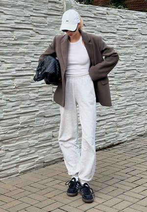 Blazer marrón de gran tamaño, camiseta sin mangas blanca, pantalones de chándal grises, bolso de mano tejido negro, gorra blanca y zapatillas oscuras. El fondo presenta una pared de piedra.