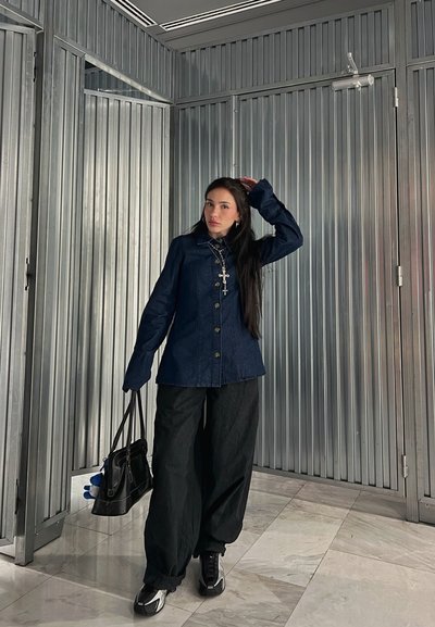 Camisa de mezclilla oscura de botones con mangas largas, combinada con pantalones anchos negros. Los accesorios incluyen un collar con cruz y un bolso negro.