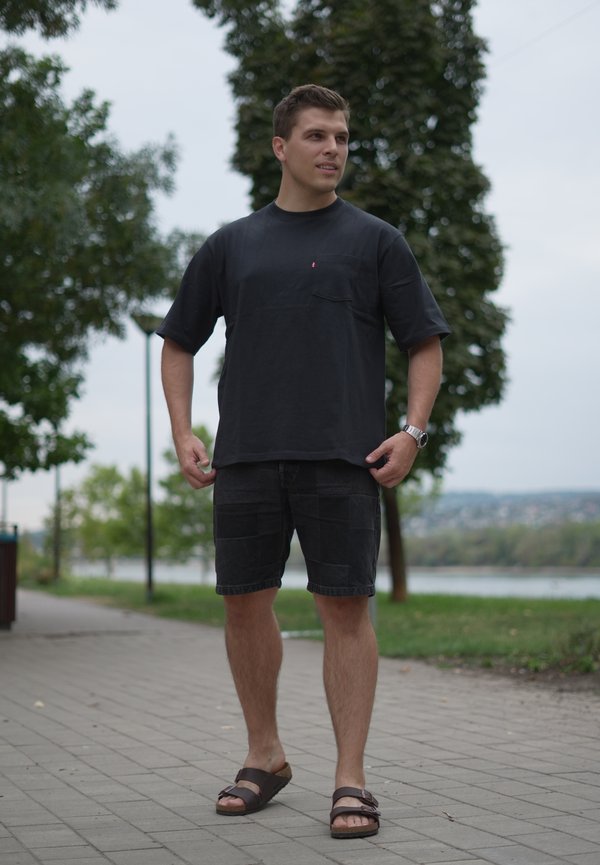 Young man wearing black t-shirt, black shorts, and sandals standing on a paved path near trees and a body of water.