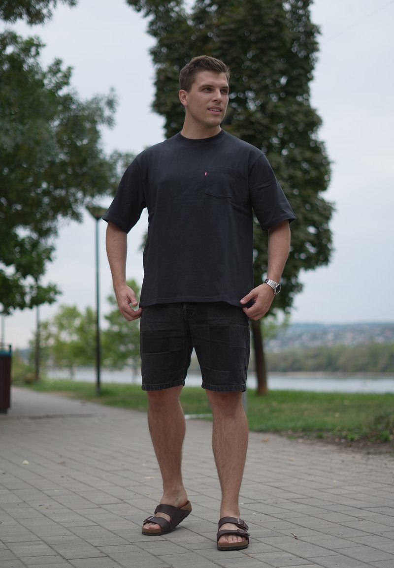Jeune homme portant un t-shirt noir, un short noir et des sandales, debout sur un sentier pavé près d'arbres et d'un plan d'eau.