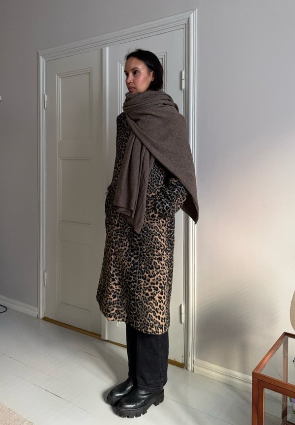 Donna in piedi all'interno che indossa un cappotto con stampa leopardata, una sciarpa marrone avvolta sulle spalle, pantaloni neri e stivali neri robusti.