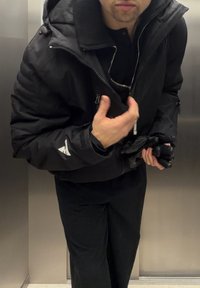 Homme portant une veste et un pantalon noirs, tenant des gants noirs, debout à l'intérieur d'un ascenseur en métal.