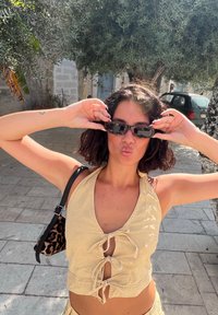 Bež halter top s vezicama na izrezu, u kombinaciji s torbom u leopardskom uzorku; kratka valovita kosa; crne sunčane naočale, vanjsko okruženje s drvećem.