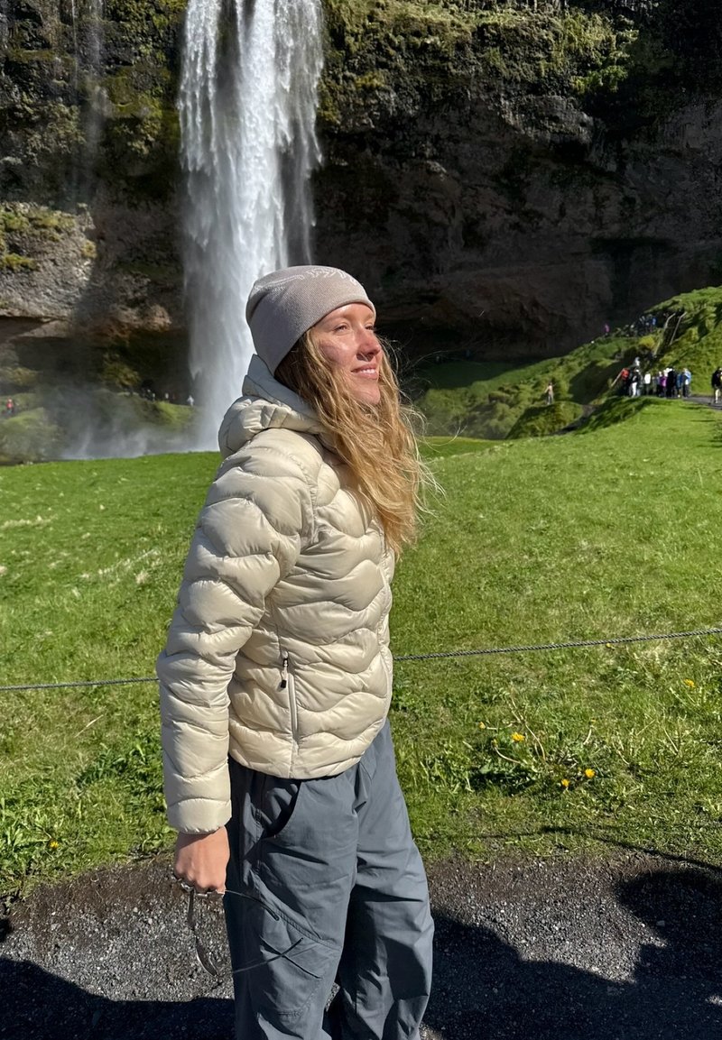 Femme en veste beige et bonnet debout près d'une cascade avec de l'herbe verte et des falaises rocheuses en arrière-plan par une journée ensoleillée.