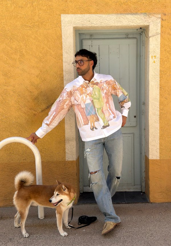 Hombre con una camisa estampada y vaqueros rotos está apoyado contra una pared amarilla, sosteniendo una correa de perro con un perro marrón claro y blanco cerca.