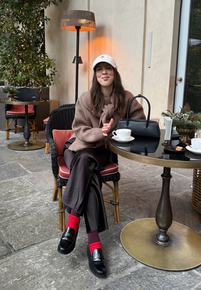 Cozy brown woollen jumper and brown satin trousers. Black handbag on a glass table with coffee cup. Red socks and black loafers. Outdoor setting.