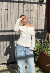 Weißer, schulterfreier Strickpullover mit geripptem Muster, kombiniert mit hellblauen, locker sitzenden Jeans, vor einem Holz-Hintergrund.
