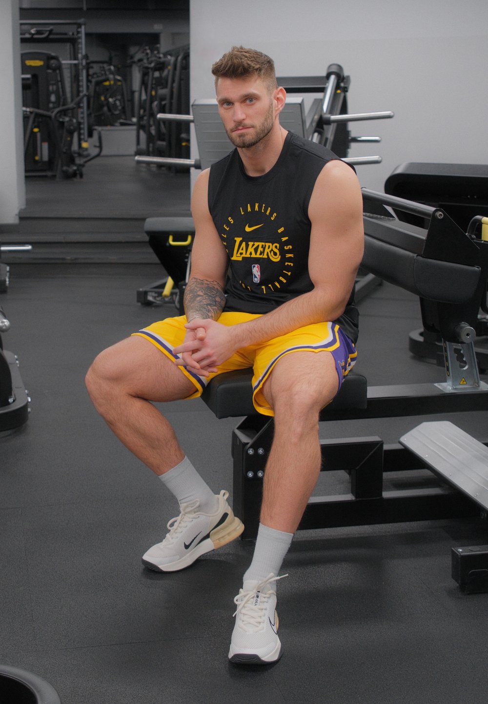 Zwart sportshirt met korte mouwen en "Lakers" logo, gele shorts met paarse accenten, zittend op een gymbank, met witte sportschoenen aan.