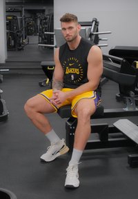 Zwart sportshirt met korte mouwen en "Lakers" logo, gele shorts met paarse accenten, zittend op een gymbank, met witte sportschoenen aan.
