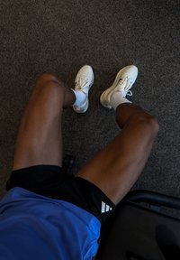 Hvide atletiksko, blå skjorte og sorte shorts vist på gulvet med et tekstureret gråt tæppe i baggrunden.