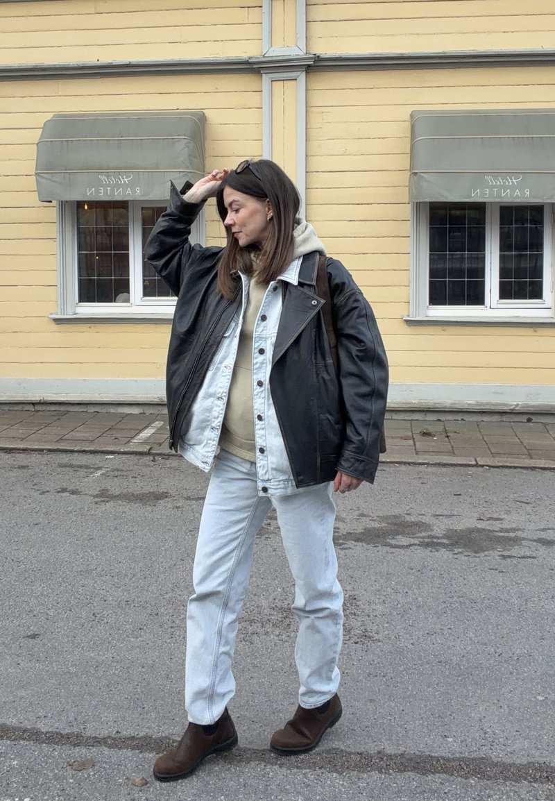 Γυναίκα που προσαρμόζει τα γυαλιά ηλίου στο κεφάλι της, φορά στρωματωτά ελαφριά τζιν και μαύρο δερμάτινο μπουφάν, μπεζ φούτερ, λευκά τζιν και καφέ μπότες στον δρόμο.