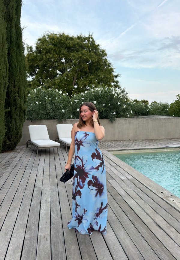 Jeune femme en robe fleurie bleue se tient sur une terrasse en bois près de la piscine, souriante et touchant ses cheveux, avec des transats et de la verdure à l'arrière.