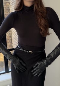 Haut à col roulé noir associé à une jupe noire et des gants en cuir. La tenue est cintrée à la taille par une ceinture noire avec une boucle dorée.
