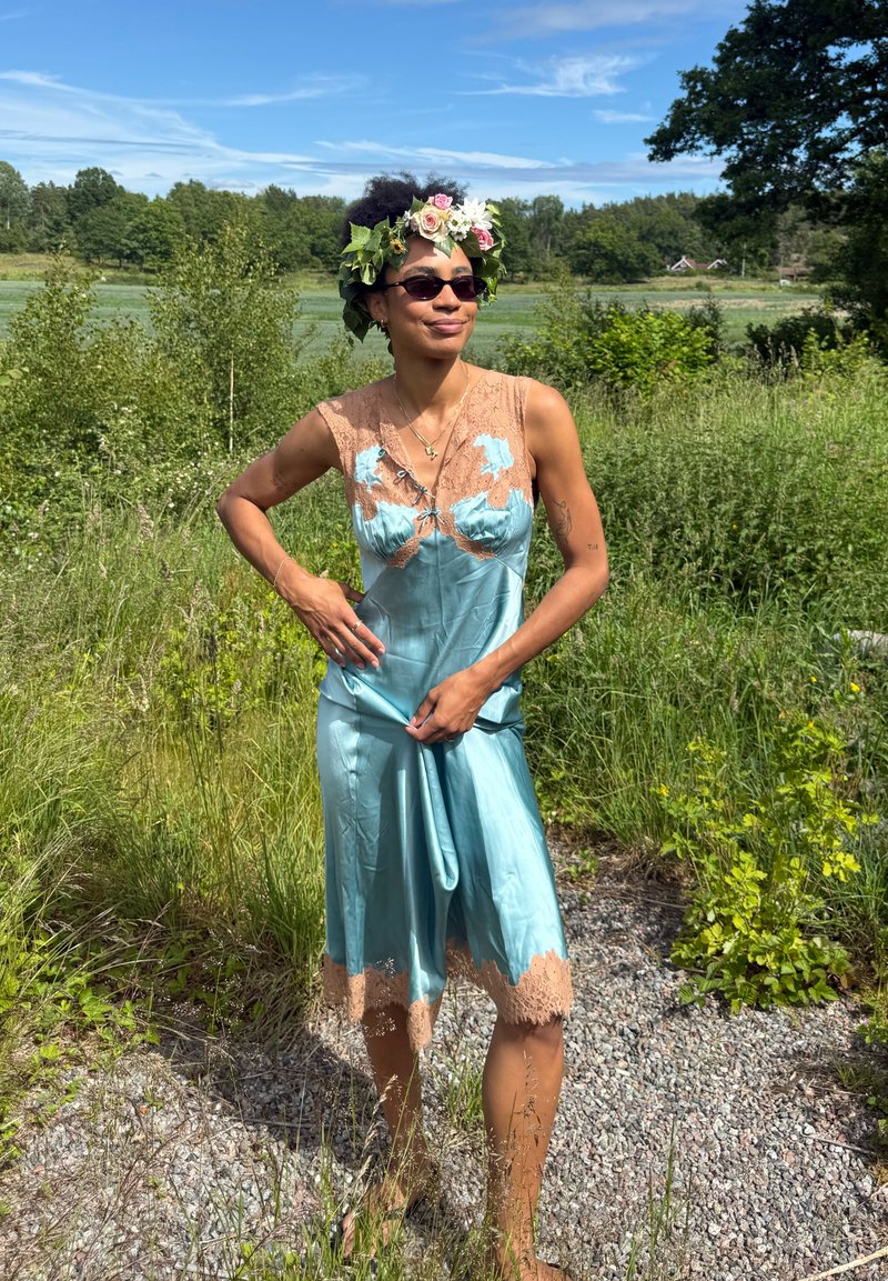 Personne portant une robe en satin bleu avec des détails en dentelle et une couronne florale, debout sur un chemin de gravier entouré d'herbe verte et d'arbres.