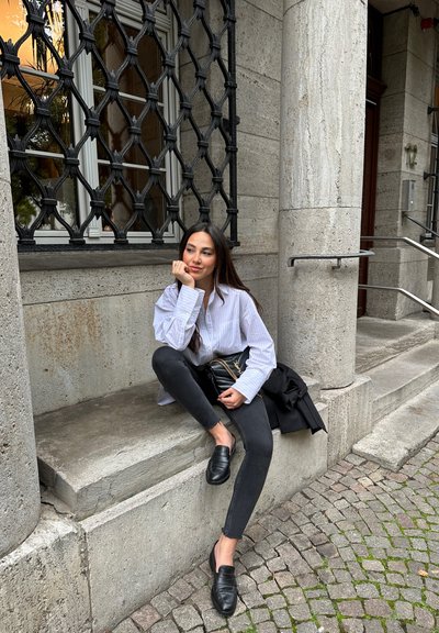 Camisa de botones blanca a rayas, pantalones vaqueros ajustados negros y mocasines negros. Sentado en un escalón de piedra con una superficie texturada y un fondo de adoquines.