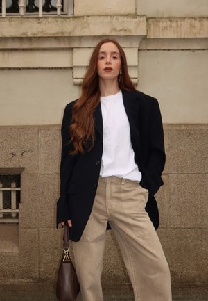 Schwarzer oversized Blazer, weißes Hemd, beiger weit geschnittener Hosen, braune Handtasche mit goldenen Akzenten, welliges Haar, vor einer strukturierten Steinmauer.