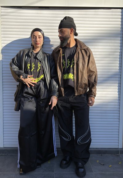 Dos personas están de pie frente a una persiana blanca, vistiendo camisetas negras con gráficos, pantalones negros holgados y chaquetas, una de cuero y otra marrón.