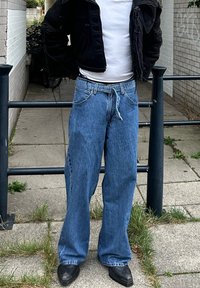 Calças de ganga largas em denim desbotado, com cintura ajustável por cordão e costura em contraste. Usadas com uma t-shirt branca e uma jaqueta preta.