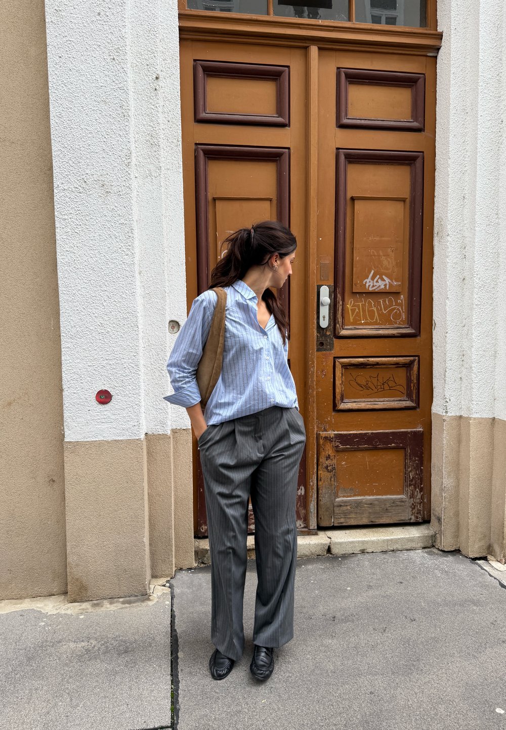 Pantalon gris à rayures fines avec une coupe décontractée, chemise à rayures bleues à boutons, et sac brun. Debout devant une porte en bois à la finition rustique.