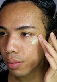 Crema applicata sulla guancia di una persona con pelle con texture, pori visibili e una leggera tonalità gialla, con uno sfondo neutro.