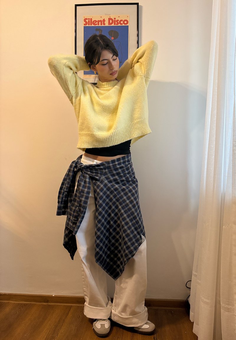 Pull court jaune, débardeur noir en dessous, chemise à carreaux bleue nouée autour d'un pantalon blanc à jambes larges, baskets grises, fond de sol en bois.
