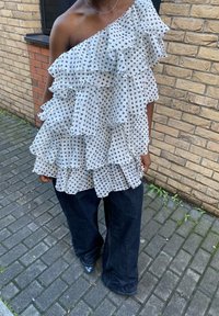 Fehér egyrészes vállpántos felső réteges fodrokkal és pöttyökkel, sötét, széles szárú farmerhez párosítva. Téglafal és járda látható a háttérben.