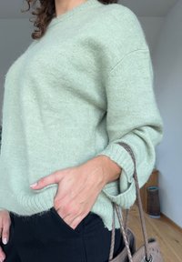 Ανοιχτό πράσινο, πλεκτό πουλόβερ με άνετη εφαρμογή, ριμπ τελείωμα και λεπτομέρειες με διπλωμένα μανίκια, συνδυασμένο με μαύρα παντελόνια και μια υφασμάτινη καφέ τσάντα.