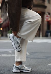 Baskets en maille grise et cuir, avec des lacets blancs et des accents noirs, présentées aux côtés d'un pantalon à jambes larges couleur crème dans une rue urbaine.