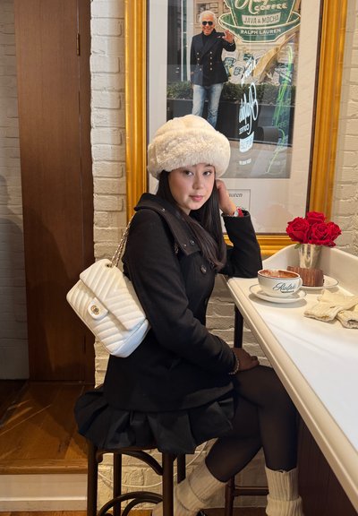 Joven mujer con ropa de invierno sentada en el mostrador de un café con café, pastel y rosas rojas junto a un póster enmarcado de Ralph Lauren detrás de ella.