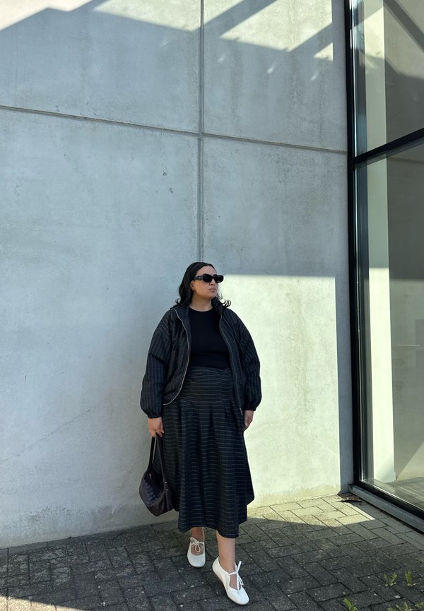 Mujer con chaqueta y falda negra a rayas, top negro, zapatos blancos, gafas de sol, sosteniendo bolso, parada junto a una pared de hormigón iluminada por el sol y una ventana de cristal.