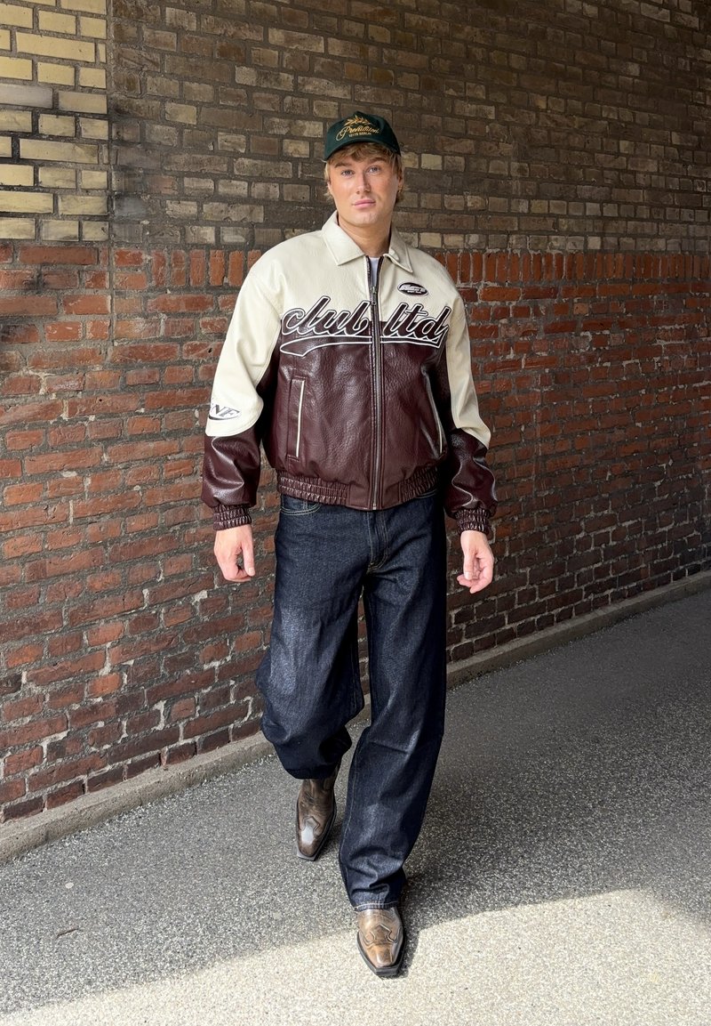 Νέος άνδρας περπατάει δίπλα σε έναν τοιχο από τούβλα φορώντας ένα δερμάτινο μπουφάν μπορντό και κρεμ, χαλαρά σκουρομπλε τζιν, καουμπόικες μπότες και μία πράσινη μπέιζμπολ κάπα.