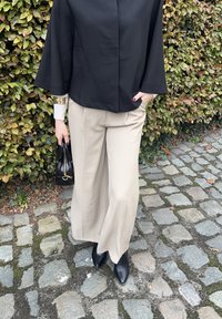 Maglietta nera a maniche corte; pantaloni larghi beige; stivali neri a punta; tiene una borsa nera con dettagli dorati; pavimentazione in pietra testurizzata.