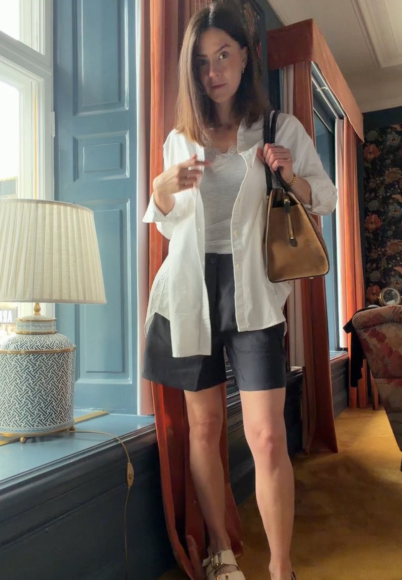 Femme portant une chemise blanche, un haut gris et un short noir, se tient près de la fenêtre tenant un sac à main marron dans une pièce avec des moulures bleues et une lampe à motifs.