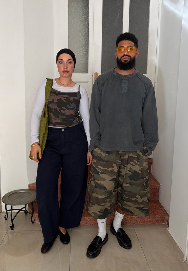 Dos adultos están dentro cerca de unos escalones; la mujer lleva una camiseta de camuflaje sobre una camisa blanca, pantalones negros y un pañuelo en la cabeza; el hombre lleva una camiseta gris, pantalones cortos de camuflaje y gafas amarillas.