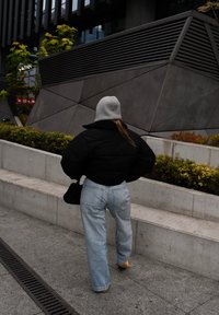 Μαύρο κοντό πλούσιο μπουφάν, ανοιχτό μπλε φαρδιά τζιν, γκρι σκούφος, μαύρη τσάντα ώμου, στέκεται κοντά σε μια υφασματική γκρίζα γεωμετρική τοιχογραφία.