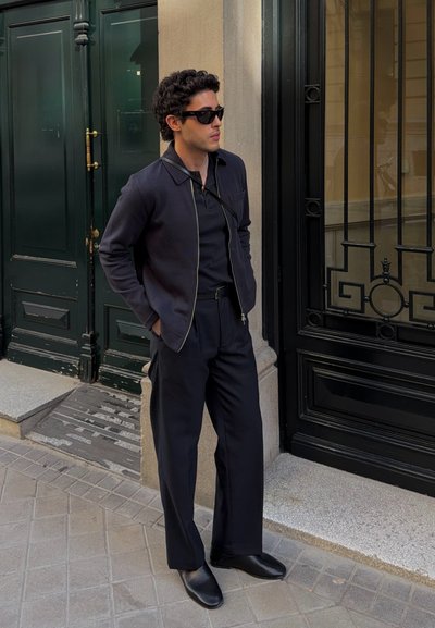 Hombre con gafas de sol negras, chaqueta oscura, polo, pantalones anchos y zapatos negros, de pie en la acera de la ciudad cerca de una puerta de hierro negra y una puerta de madera verde.