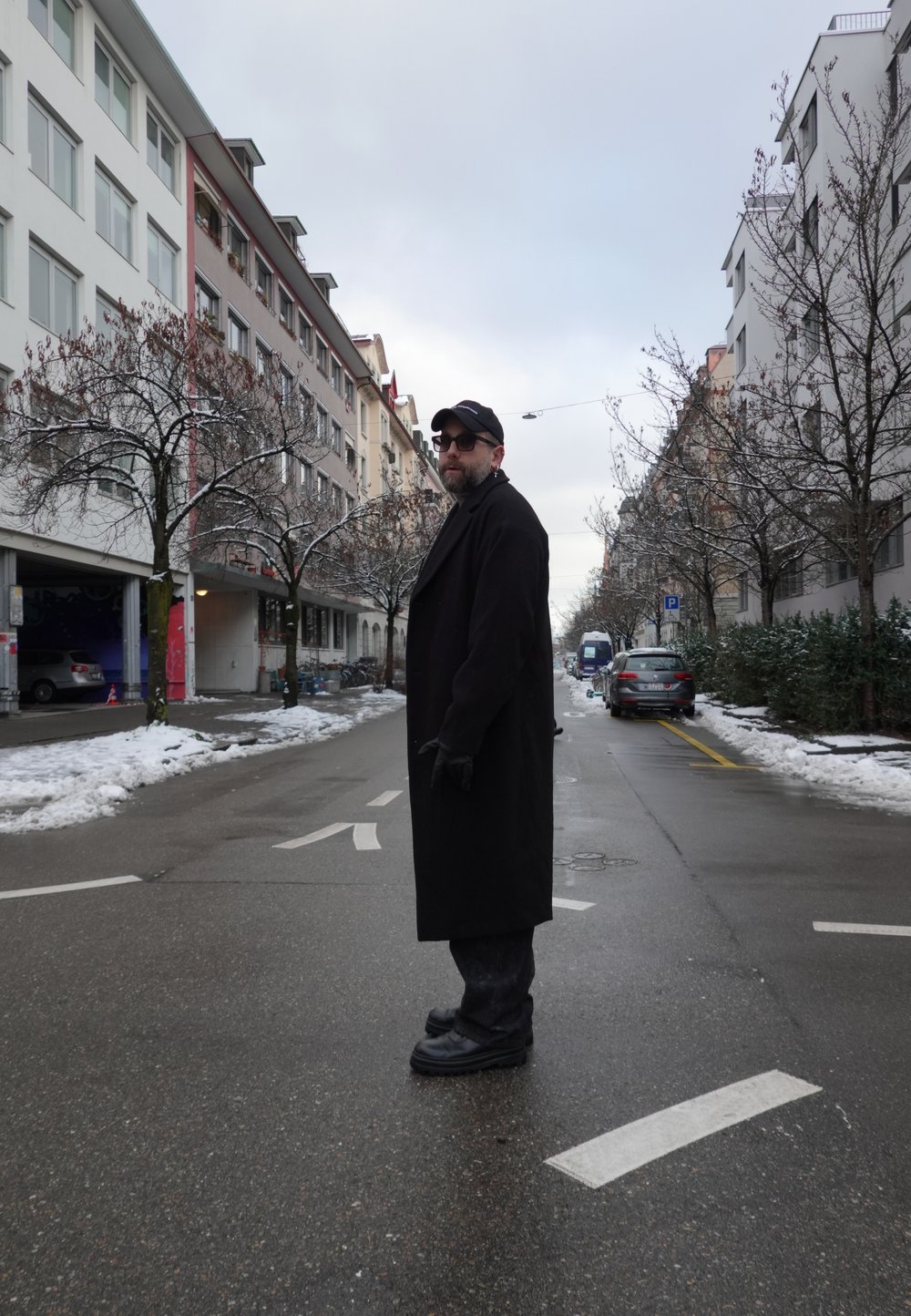 Homme portant un manteau noir, des gants, une casquette et des lunettes de soleil, debout dans une rue mouillée avec des plaques de neige, entouré d'arbres nus et d'immeubles d'appartements.