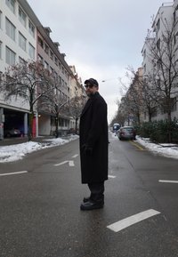 Homme portant un manteau noir, des gants, une casquette et des lunettes de soleil, debout dans une rue mouillée avec des plaques de neige, entouré d'arbres nus et d'immeubles d'appartements.