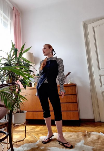 Chaleco negro sobre una blusa a cuadros con mangas abullonadas, combinado con pantalones negros cortos y sandalias de tacón; ambiente interior con plantas.
