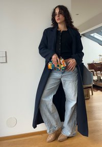 Manteau long bleu marine sur un haut noir, jean bleu clair à jambes larges, et chaussures pointues couleur nude. Tient un sac pochette à motifs colorés.